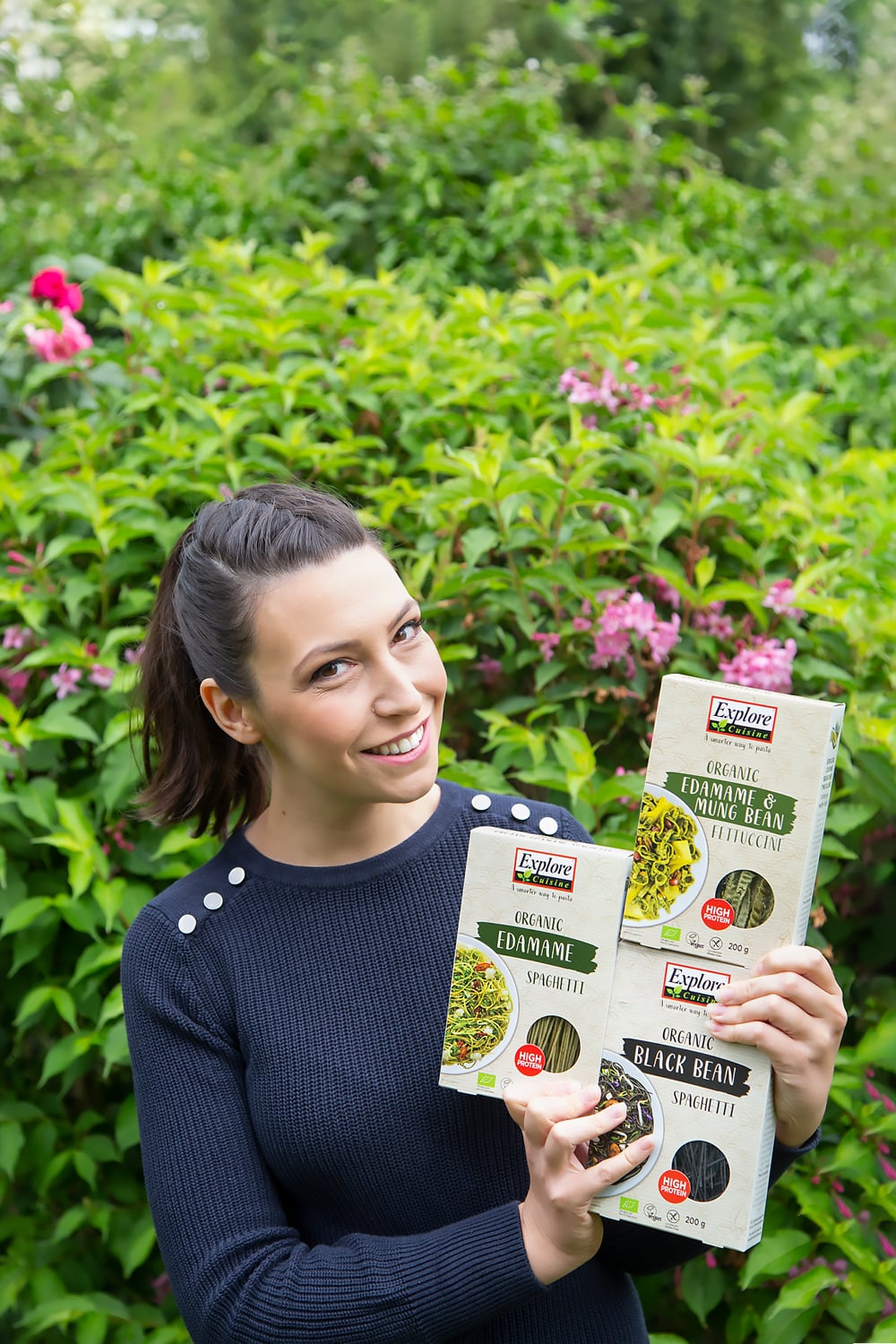 Holding three packs of Exlpore Cuisine Products; Organic Edamame and Mung Bean Fettuccini, Organic Edamame Spaghetti, Organic Black Bean Spaghetti