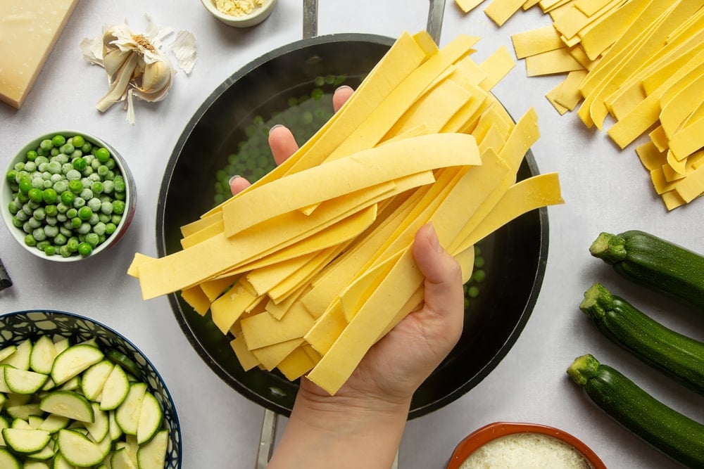 Lasagne sheets cut into strips, being added to boiling water with peas.