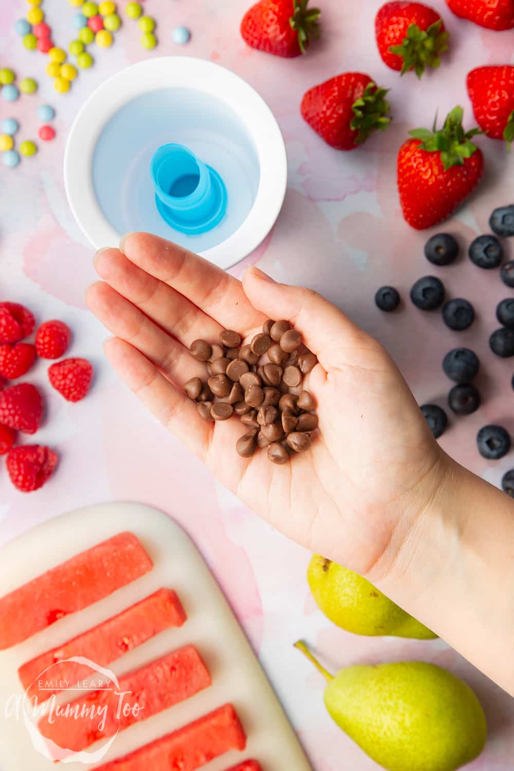 YoPop frozen yogurt maker open with a hand holding small pieces of chocolate with more fruit in the background.