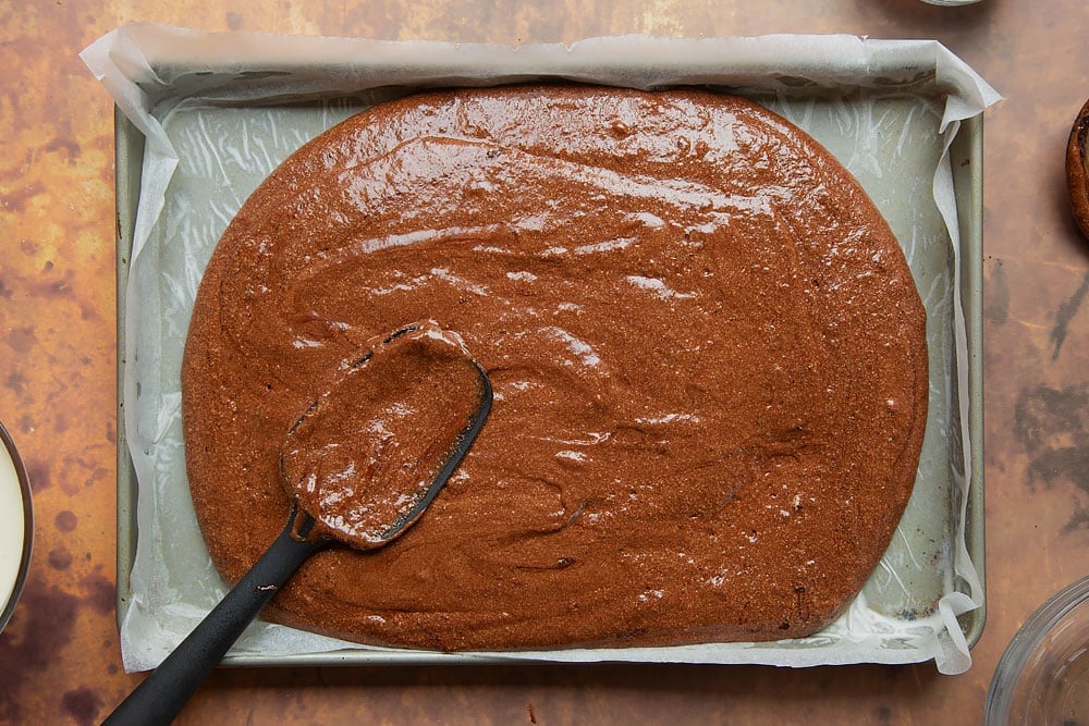 Chocolate roulade batter poured into lined swiss roll tin - silicone spatula beginning to level the batter off.