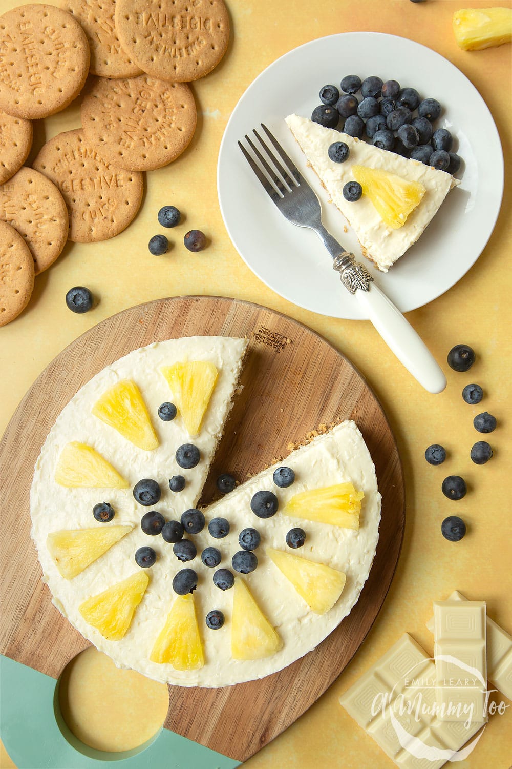 Extra special no-bake coconut cheesecake topped with pineapple pieces and blueberries. Served to a board with a slice on a plate.