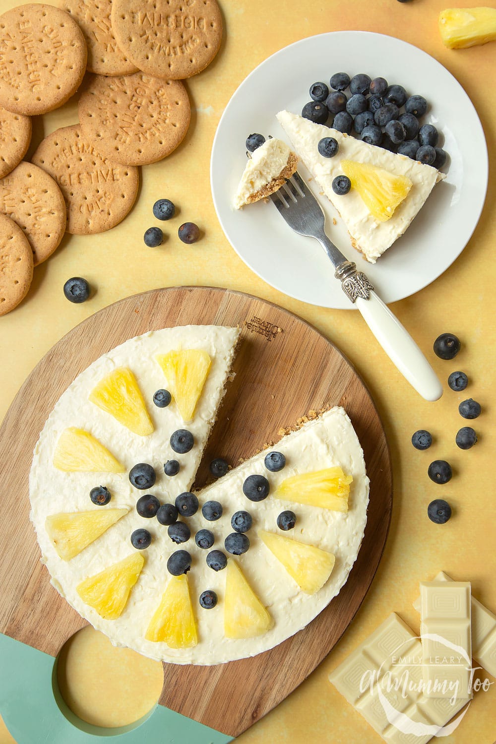 Extra special no-bake coconut cheesecake, decorated with fruit, served on a serving board. One slice is served to a small plate with blueberries and a fork.