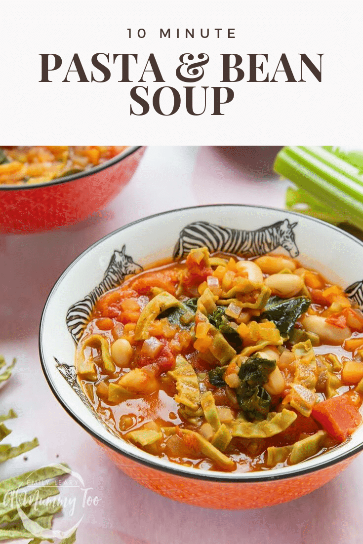 A bowl of hot pasta e fagioli with all the ingredients peeking out the sauce. Some raw ingredients are visible on the table.