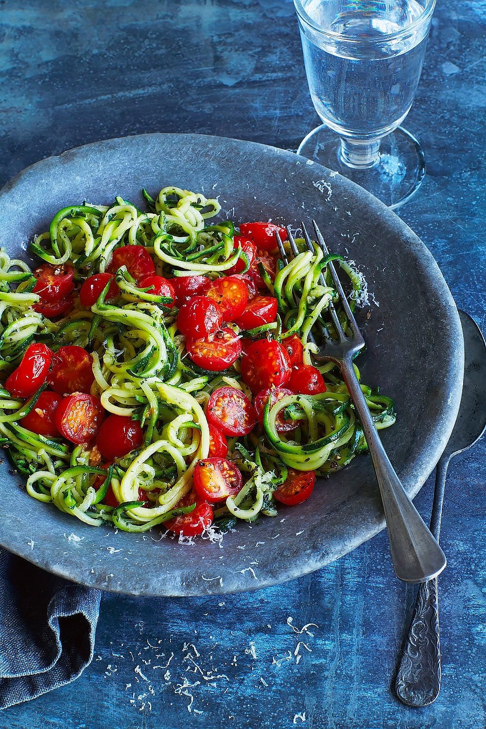 Annabel Karmel's courgette spaghetti, served into a bowl with a fork and spoon and a glass of water.