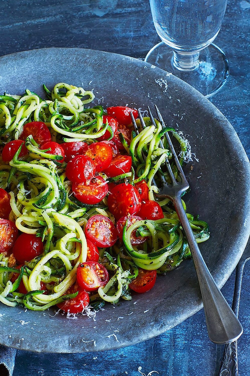 Annabel Karmel's courgette spaghetti, served into a bowl with a fork and a glass of water. Close up