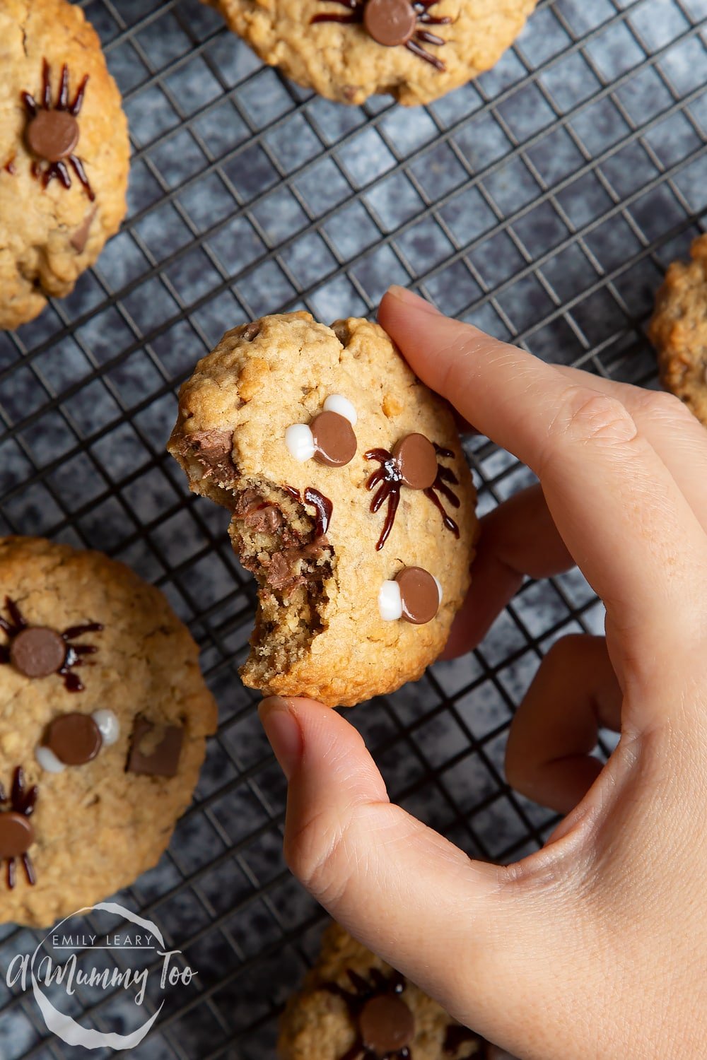 Halloween peanut butter spider cookies cooling on a wire rack. A hand is holding one cookie, with a bite in the cookie revealing chocolate chips inside.