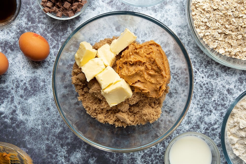 A bowl containing sugar, butter and peanut butter. Surrounded by ingredients for Halloween peanut butter spider cookies