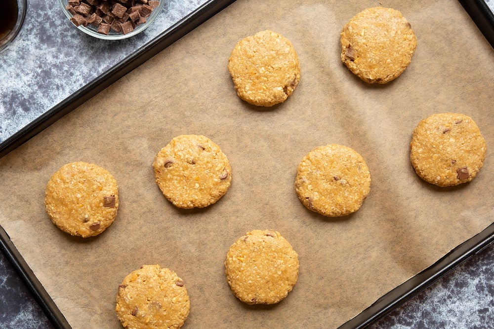 Eight raw Halloween peanut butter spider cookies on a baking sheet lined with baking paper. They have been pressed down to form discs.