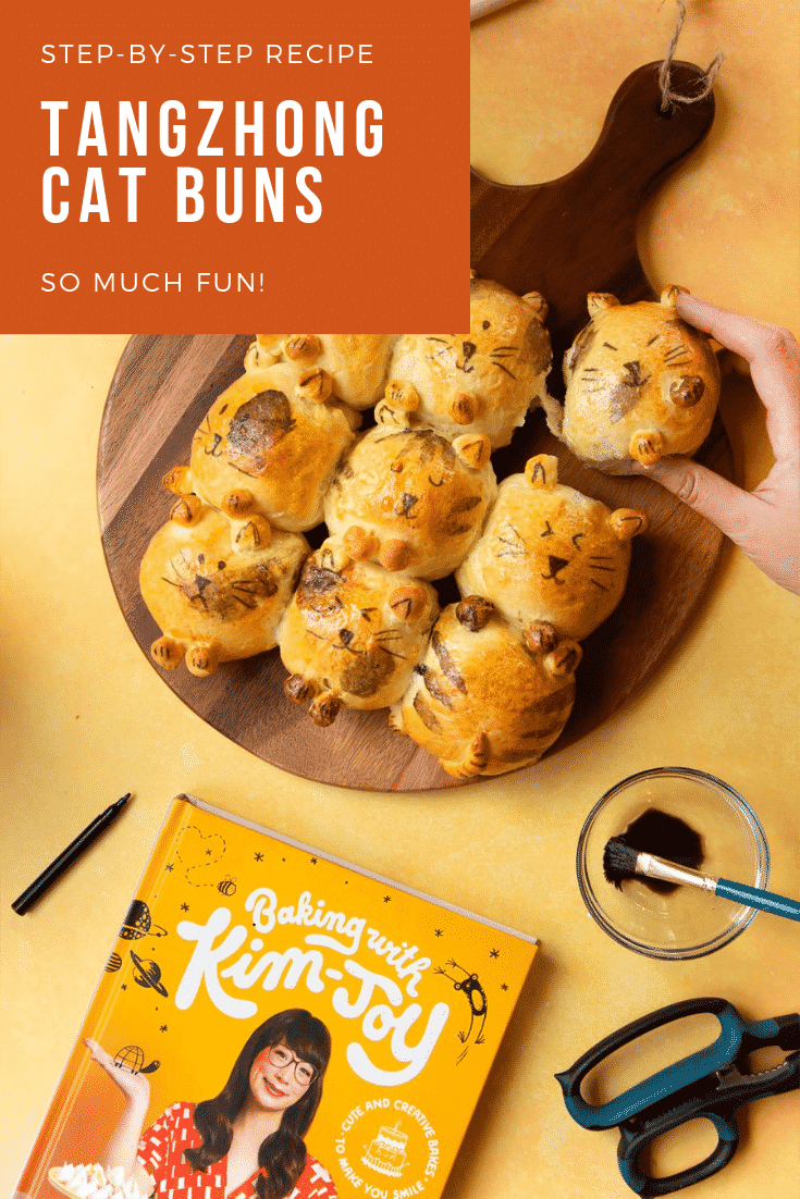 A set of 9 tangzhong bread buns shaped to look like kittens. Arranged on a dark bread board, with a hand tearing one bun away.