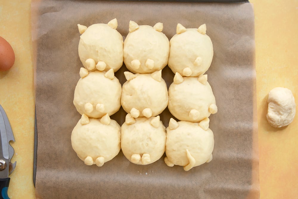 9 balls on tangzhong bread dough arranged on a tray lined with baking paper. Cats ears, feet and tails made from dough and attached to the rolls.