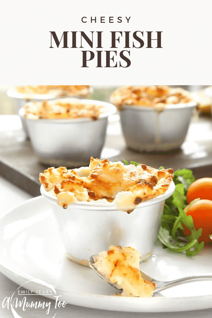 A spoon sits on a white plate topped with mini cheesy fish pie. The pie itself sits on a white plate with a side of salad. In the background there's some additional mini fish pies on a wooden serving board. At the top of the image there's some text describing the image for Pinterest. 