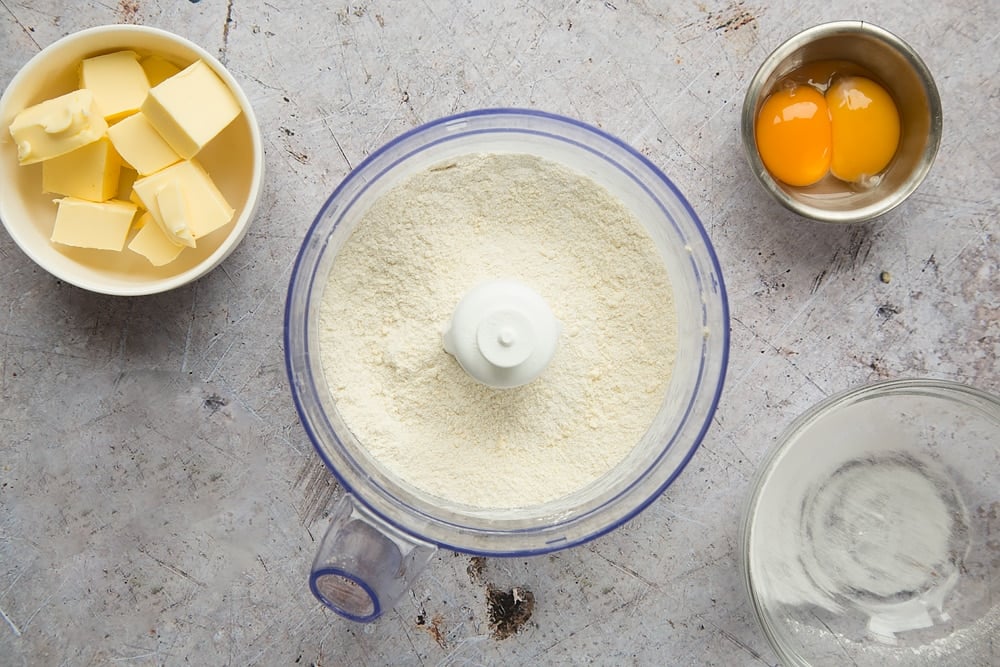 Butter crumbled flour mixture in a blender bowl with 2 other bowls holding butter and eggs.