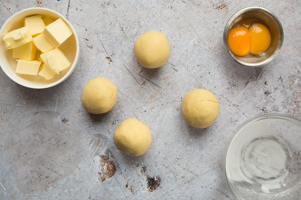 yellow like coloured pastry dough balls on a surface accompanied by butter and egg yolks in clear bowls.