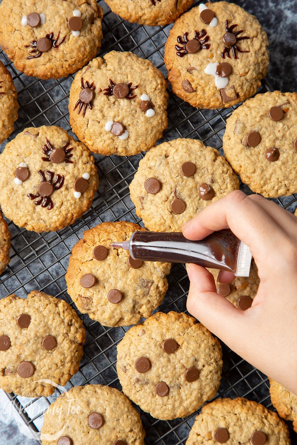 Halloween peanut butter spider cookies on a cooling rack. A hand is using a dark brown icing pen to add legs to the chocolate spiders.