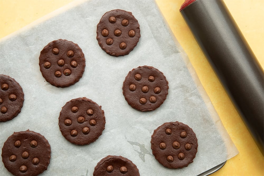 Chocolate shortbread cookie dough rounds placed on a tray lined with baking paper. Each cookie has a few chocolate drops pressed into its surface.