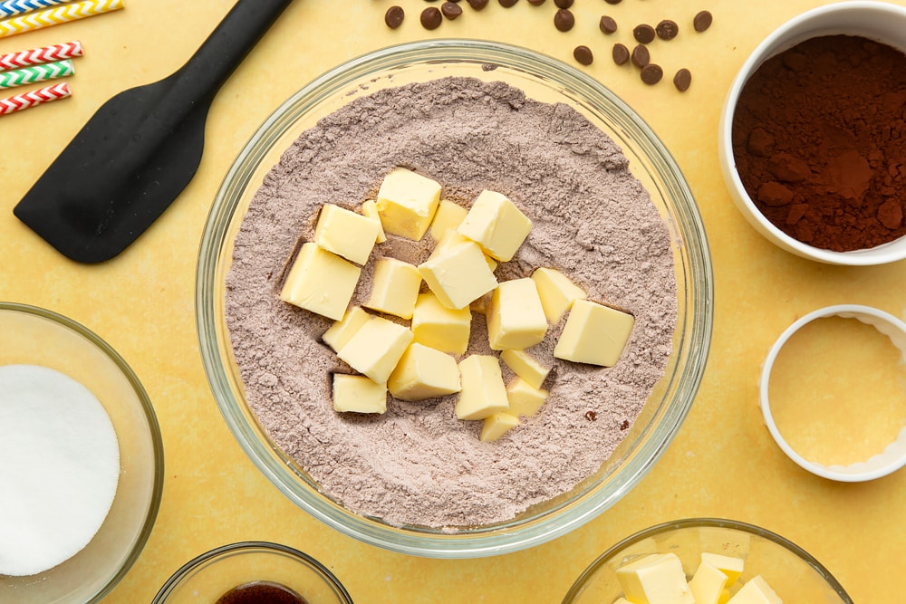 A mixing bowl containing flour, sugar and cocoa mixed together, cubed butter on top. Ingredients for chocolate shortbread arrange around the outside.