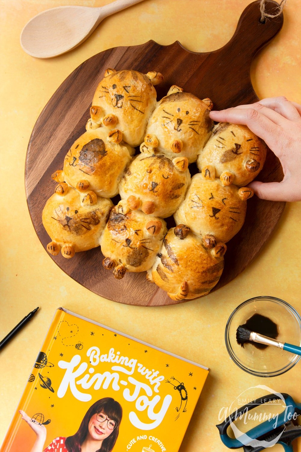 Kim-Joy's Tangzhong Cat Buns - nine bread rolls on a board, shaped and decorated to resemble cats, a hand reaches for one bun.