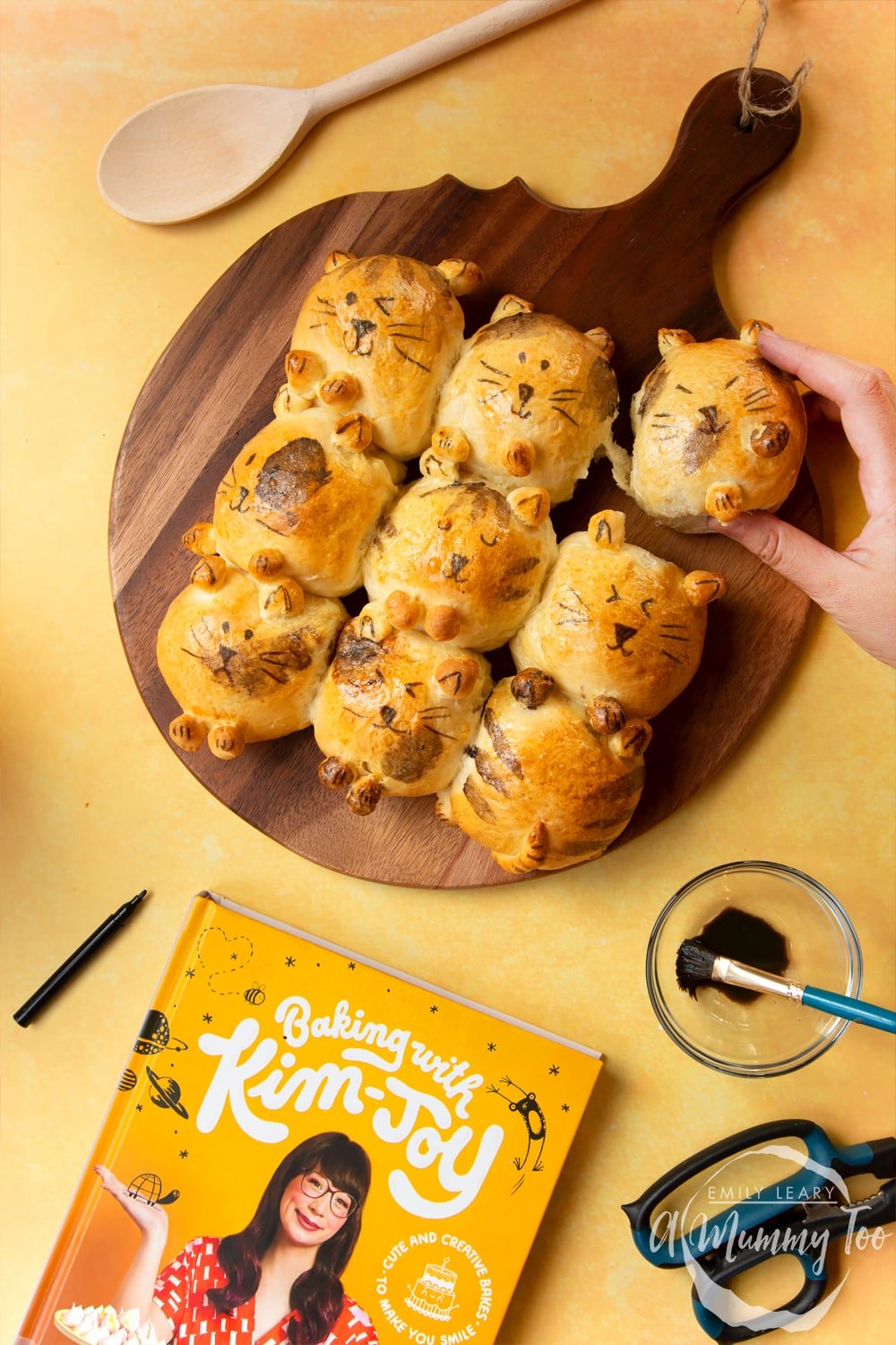 Kim-Joy's Tangzhong Cat Buns - nine bread rolls on a board, shaped and decorated to resemble cats A hand tears away one bun.