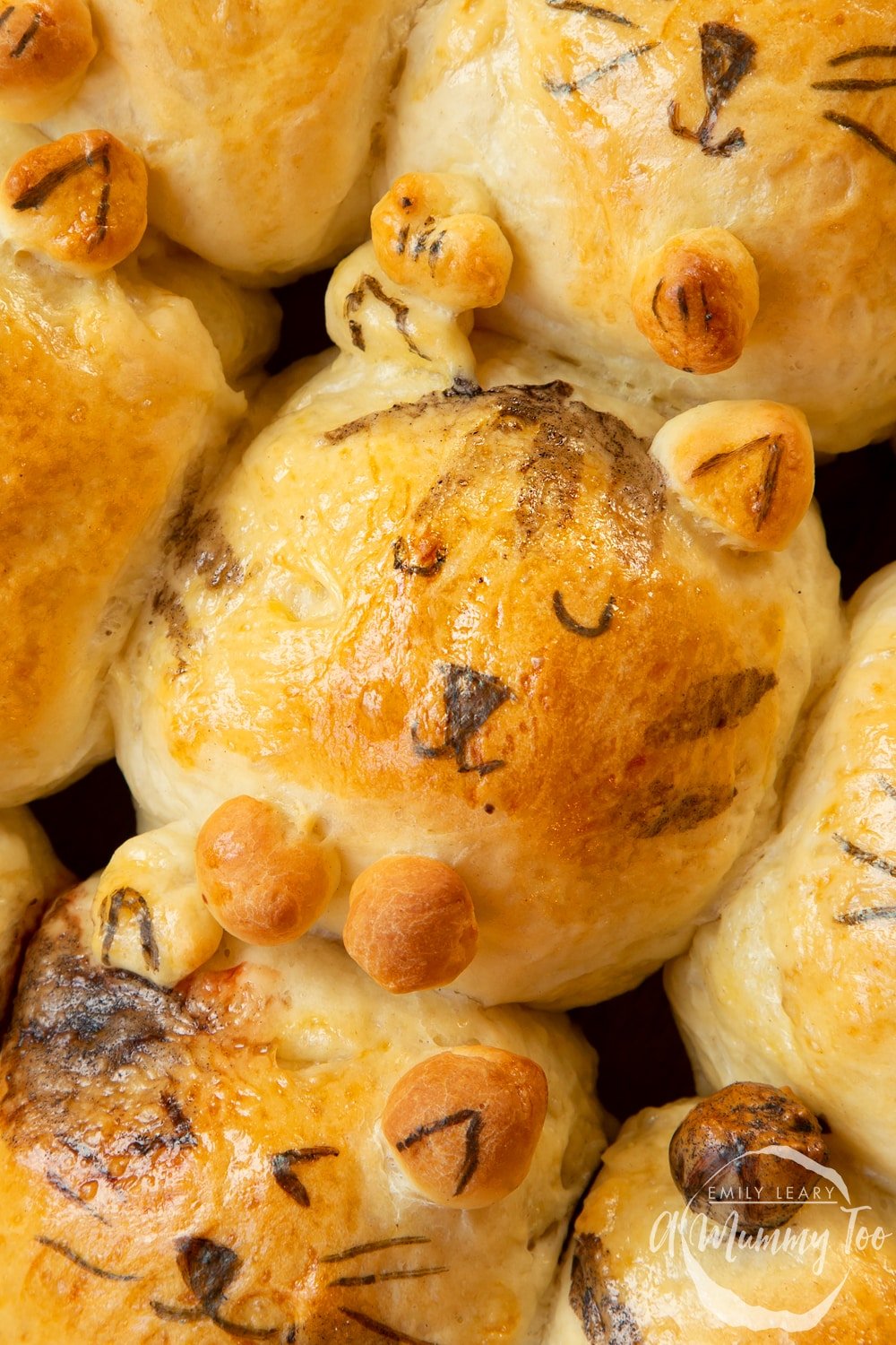 9 cat shaped tangzhong bread rolls on a tray lined with baking paper. Baked and cooling on a wire rack. Cat fur patterns painted on with food colouring. Facial features added with a felt tip. 