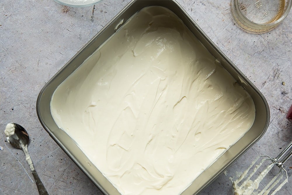 "Overhead shot 
 of cream cheese mixture in a square loose-bottomed straight-sided tin surrounded by a mixer, jar, and spoon"
