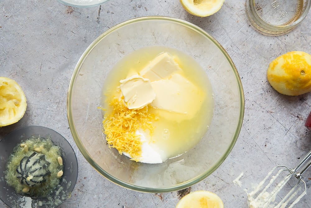 "Overhead shot 
 of lemon juice, lemon rind, butter and sugar in a clear bowl surrounded by lemons, jar, mixer, and lemon squeezer"