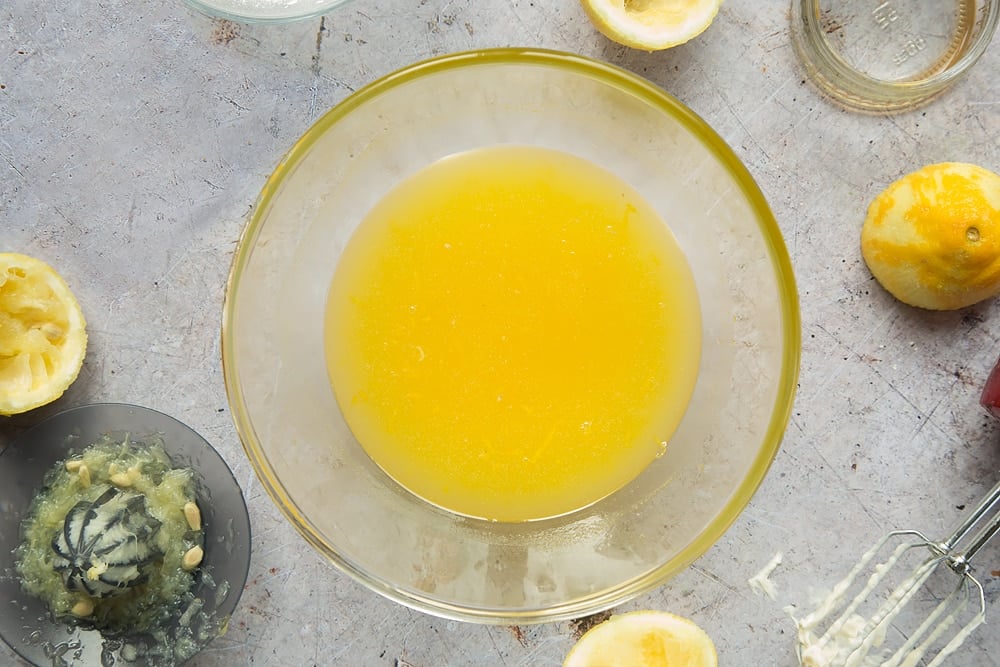 Overhead shot of melted butter in a clear bowl surrounded by lemons, jar, mixer, and lemon squeezer