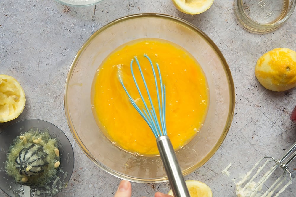 Overhead shot of whisked eggs with whisk in a clear bowl surrounded by lemons, jar, mixer, and lemon squeezer