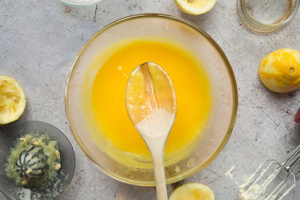 Overhead shot of wooden spoon below egg mixture in a clear bowl surrounded by lemons, jar, mixer, and lemon squeezer