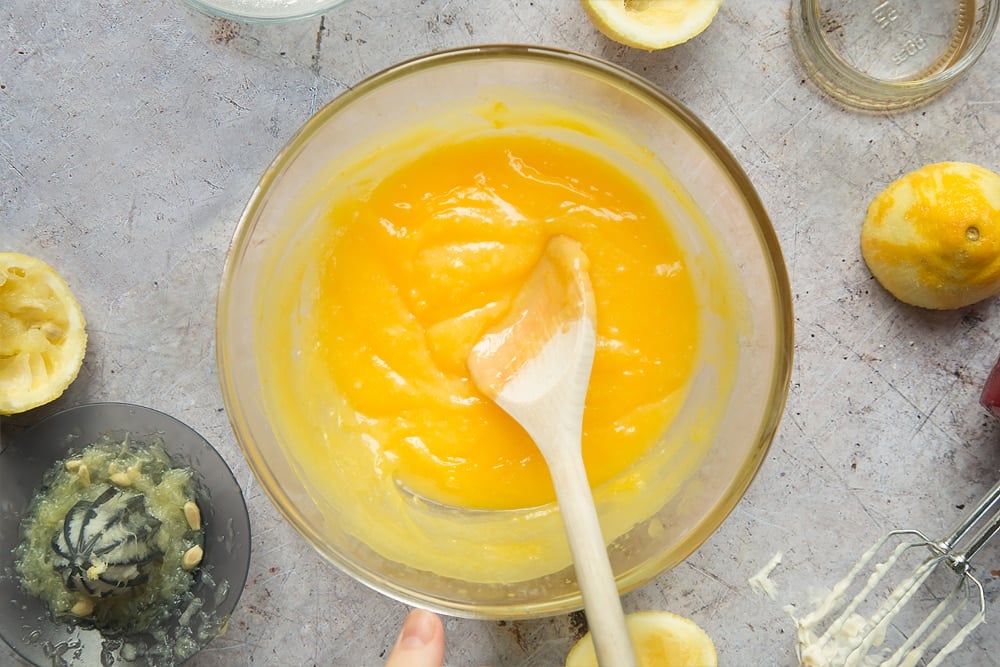 Overhead shot of wooden spoon in egg mixture in a clear bowl surrounded by lemons, jar, mixer, and lemon squeezer