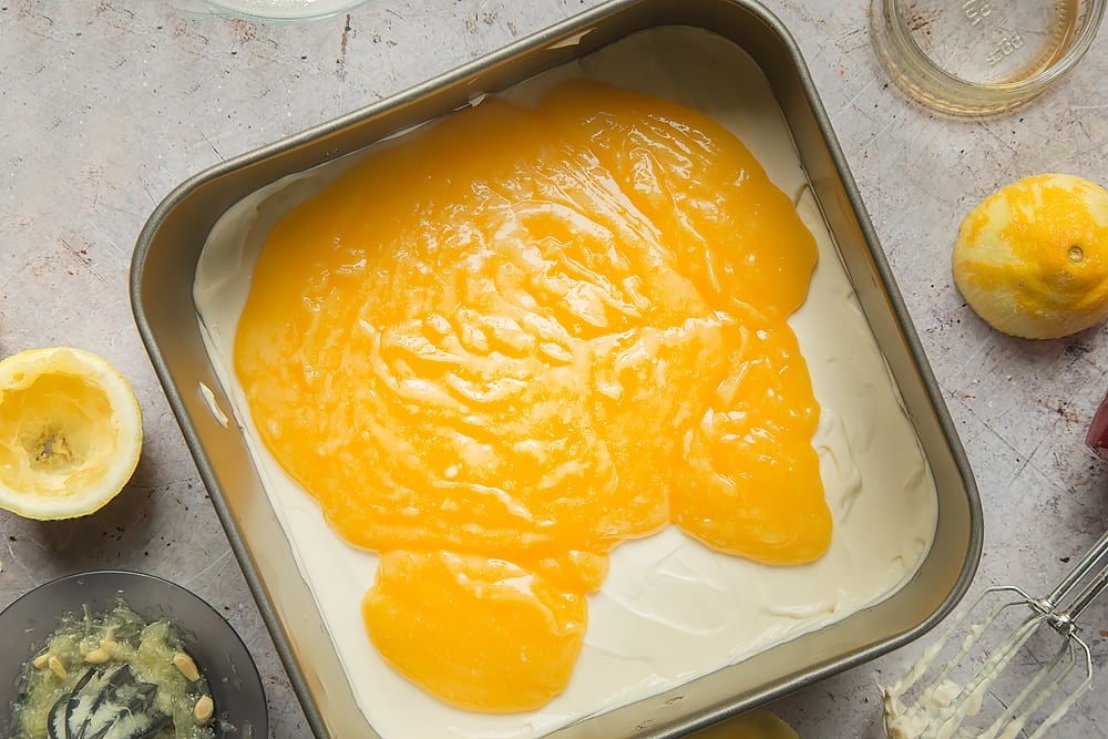 Overhead shot of cream cheese filling topped with lemon curd in a square tin surrounded by lemons, jar, mixer, and lemon squeezer