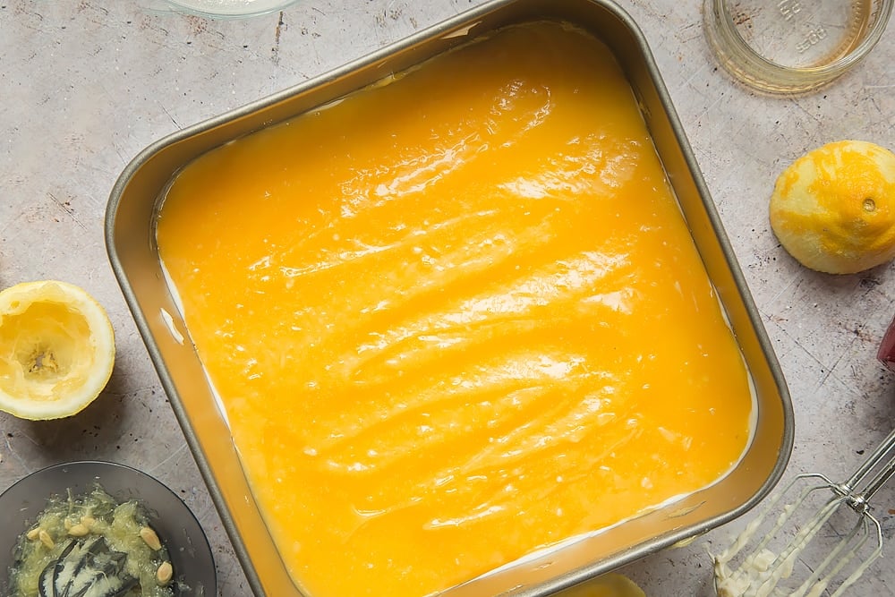 Overhead shot of Lemon curd yogurt cheesecake in a square tin surrounded by lemons, jar, mixer, and lemon squeezer