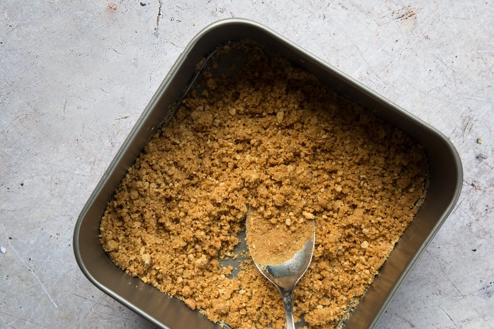 Overhead shot of digestive biscuits crumbs with spoon in a square loose-bottomed straight-sided tin