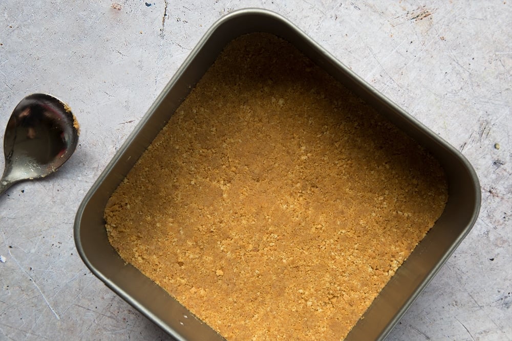 Overhead shot of digestive biscuits crust in a square loose-bottomed straight-sided tin and a spoon on the side