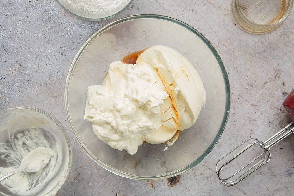 Overhead shot of cream cheese, yogurt, and vanilla in a clear bowl surrounded by a mixer and empty bowl