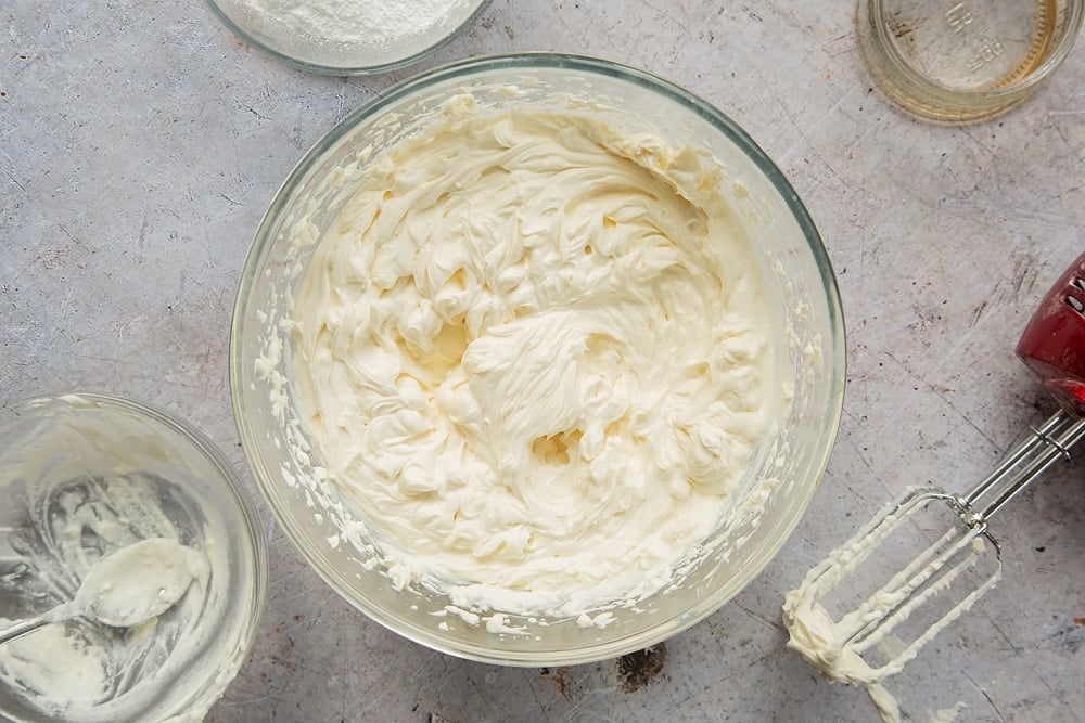 Overhead shot of whisked cream cheese, yogurt and vanilla in a clear bowl surrounded by a mixer and empty bowl