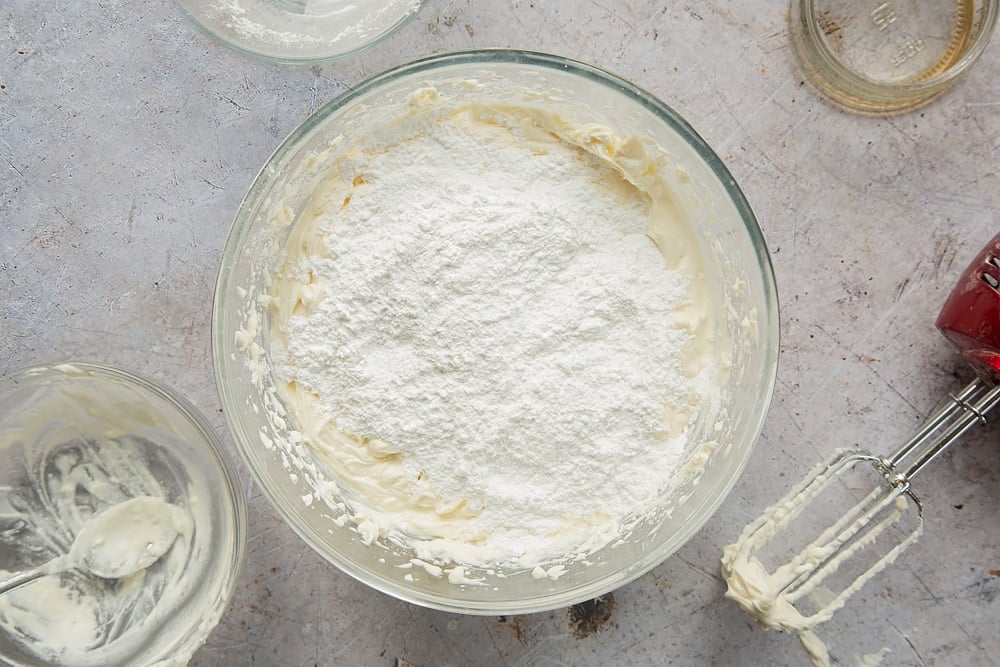 Overhead shot of sugar on top of cream cheese mixture in a clear bowl surrounded by a mixer and empty bowl