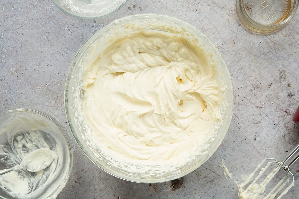 Overhead shot of whisked cream cheese, yogurt and vanilla in a clear bowl surrounded by a mixer and empty bowl