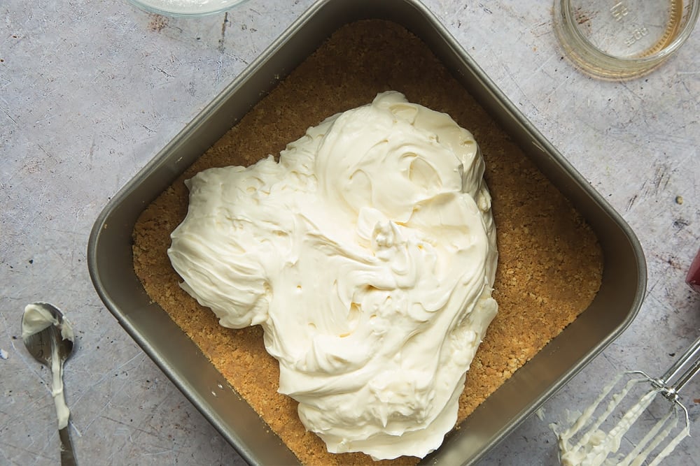 Overhead shot of cream cheese mixture on top of digestive biscuits crust in a square loose-bottomed straight-sided tin surrounded by a mixer, jar, and spoon