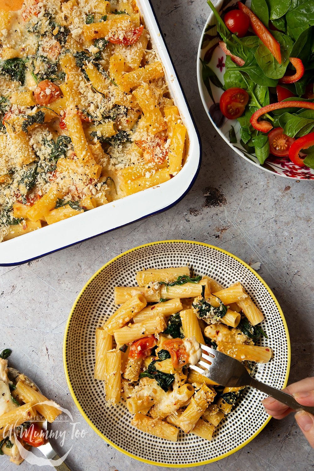 A fork going into the bowl of Cherry tomato, spinach and garlic mozzarella pasta bake. The bowl is sat alongside the pan with the remainder of the Cherry tomato, spinach and garlic mozzarella pasta bake alongside a side salad. 