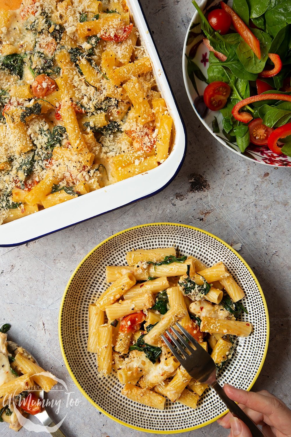 Overhead shot of a decorative bowl with a serving of Cherry tomato, spinach and garlic mozzarella pasta bake alongside the pan and a side salad. 