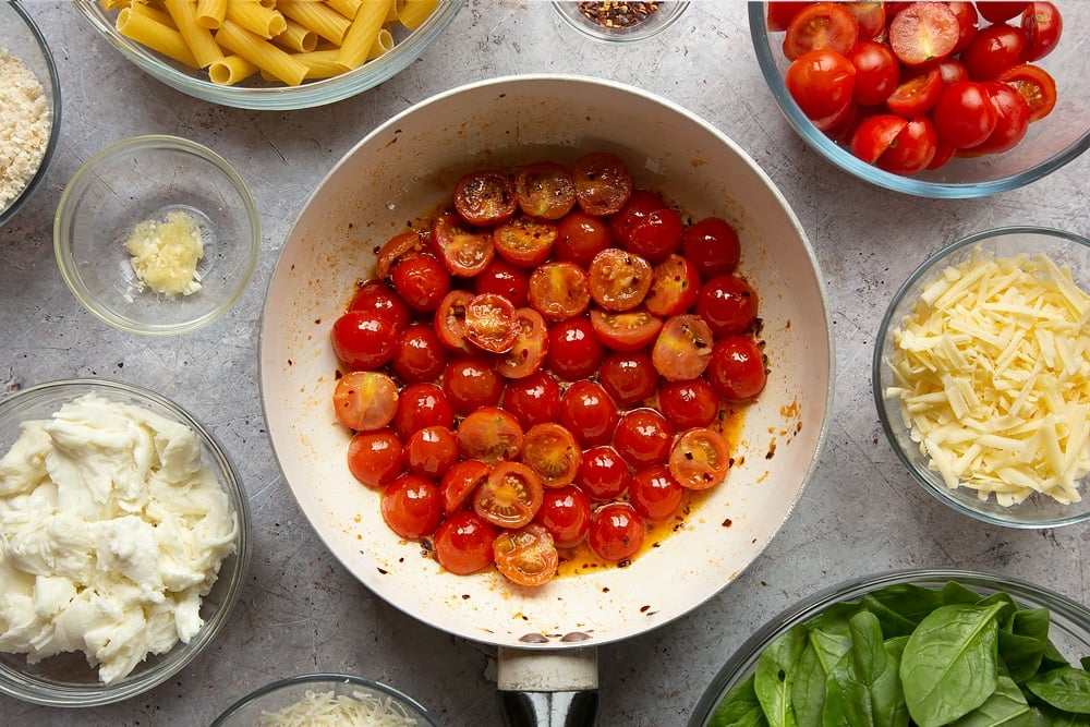 Chopped cherry tomatoes in the pan having been cooked. The shot is taken overhead and the pan is surrounded by ingredients required to make the Butter melting in a pan surrounded by different ingredients required to make Cherry tomato, spinach and garlic mozzarella pasta bake. 