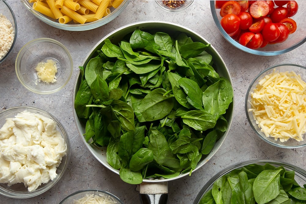 Overhead shot of spinach in a pan surrounded by ingredients required to make the Butter melting in a pan surrounded by different ingredients required to make Cherry tomato, spinach and garlic mozzarella pasta bake. 