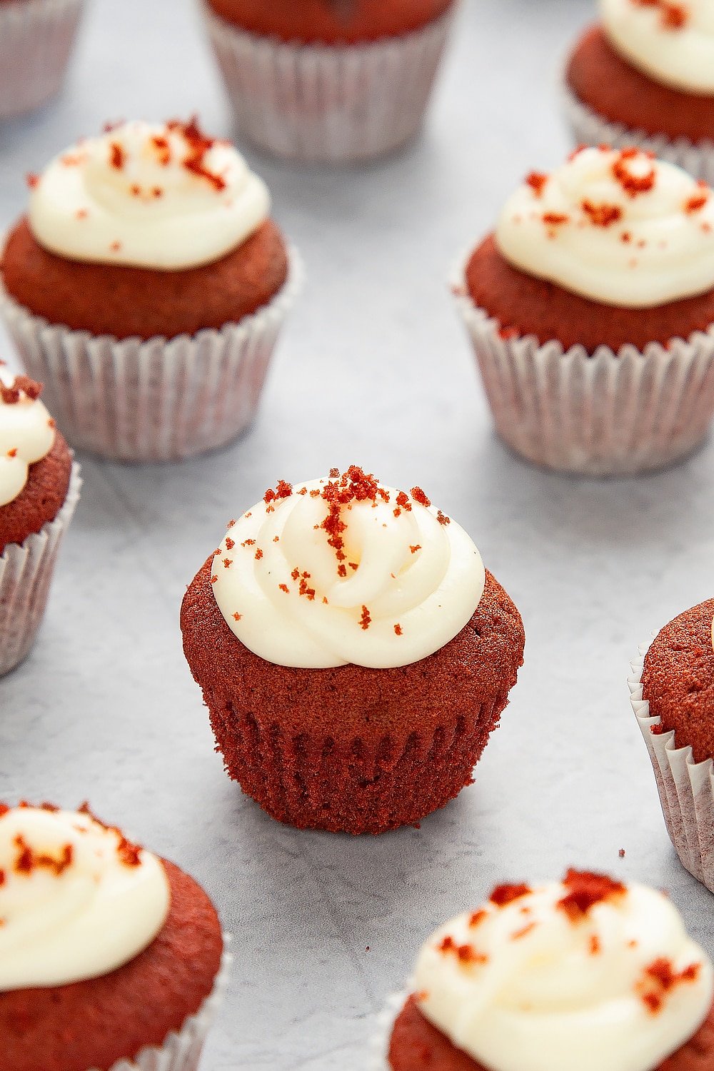 Mini red velvet cupcakes with cream cheese frosting unwrapped from it's case sat on a grey table top surrounded by additional cupcakes.