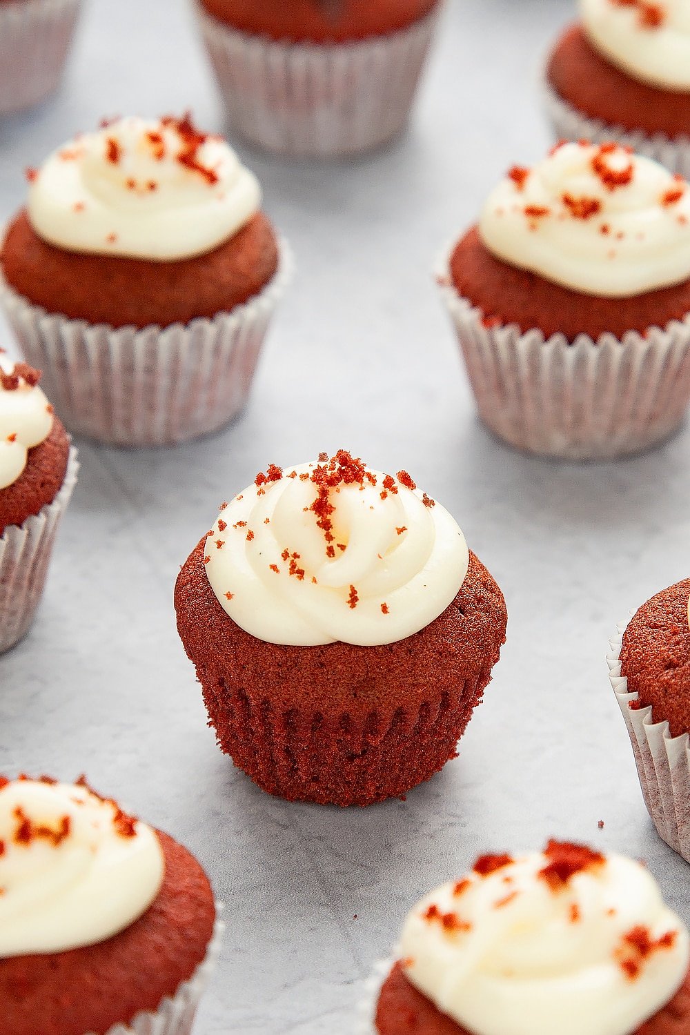 Mini red velvet cupcakes with cream cheese frosting unwrapped from it's case sat on a grey table top surrounded by additional cupcakes. 