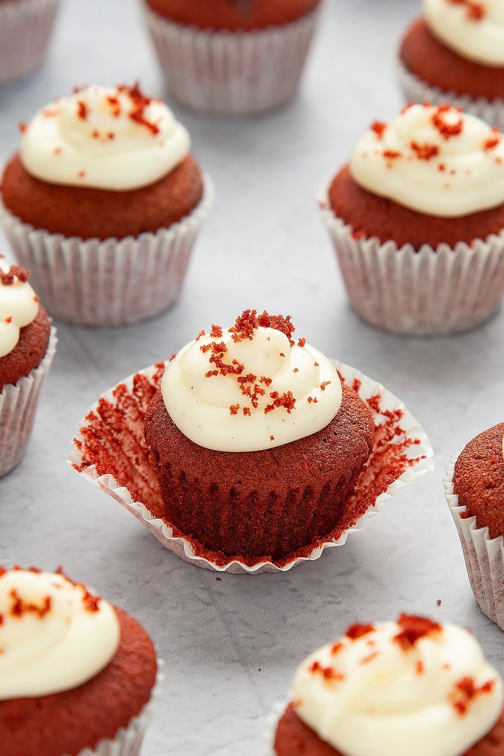 mini red velvet cupcakes with cream cheese frosting being unwrapped from the wrapper surrounded by additional mini red velvet cupcakes with cream cheese frostings on a grey table.