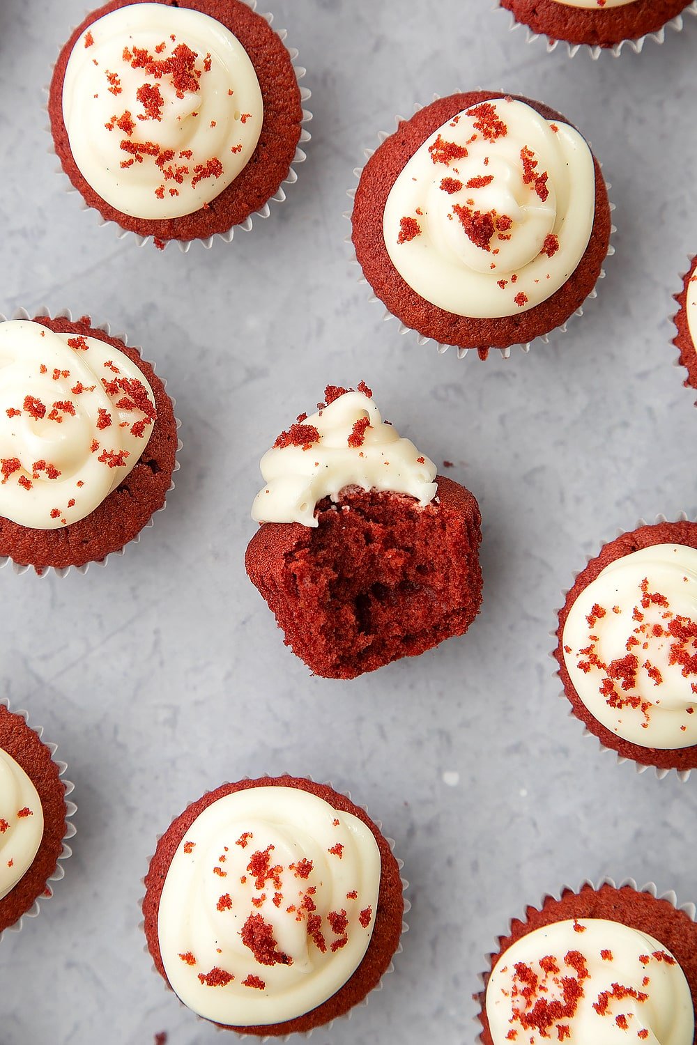 Overhead shot of mini red velvet cupcakes with cream cheese frosting. The centred cupcate is sat on its side with a bite taken out. 