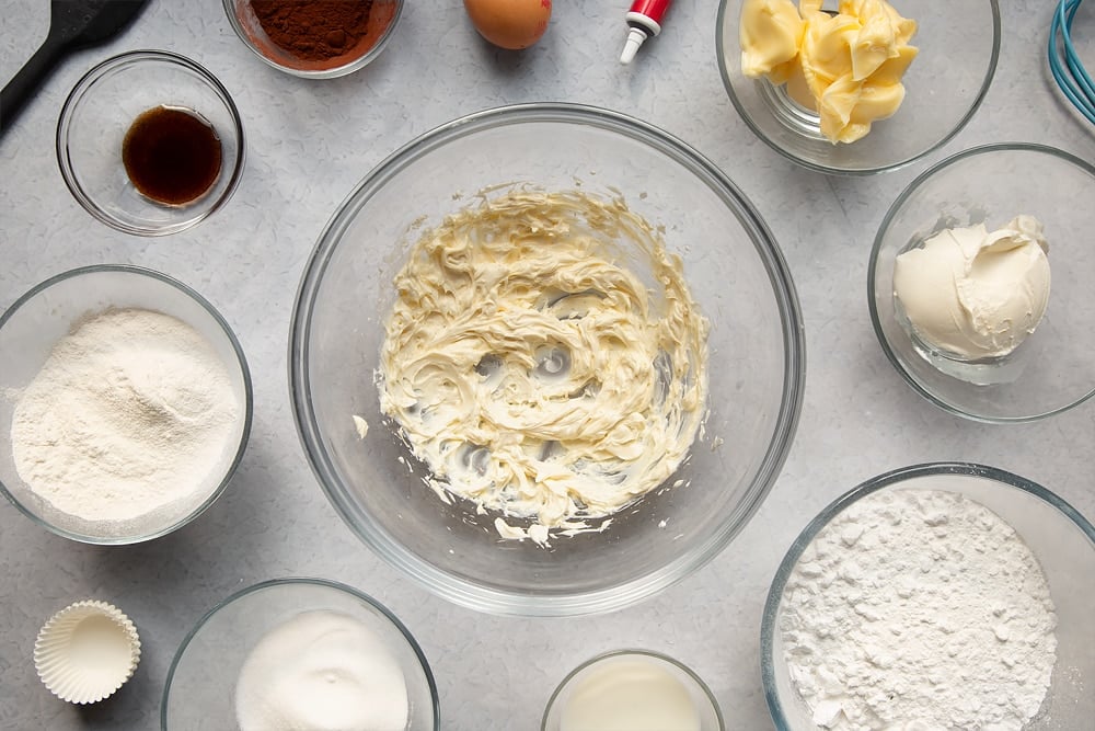 Overhead shot of a bowl of ingredients for the cream cheese frosting being mixed togethered surrounded by additional ingredients which are required for the recipe. 