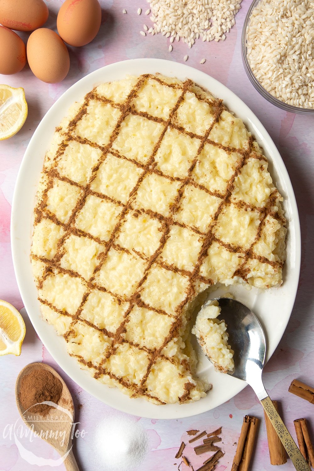Arroz Doce (Portuguese rice pudding) on a large, white, oval-shaped plate. The rice pudding is decorated with ground cinnamon in a crosshatch pattern. A spoon rests on the plate where some of the pudding has been taken away.