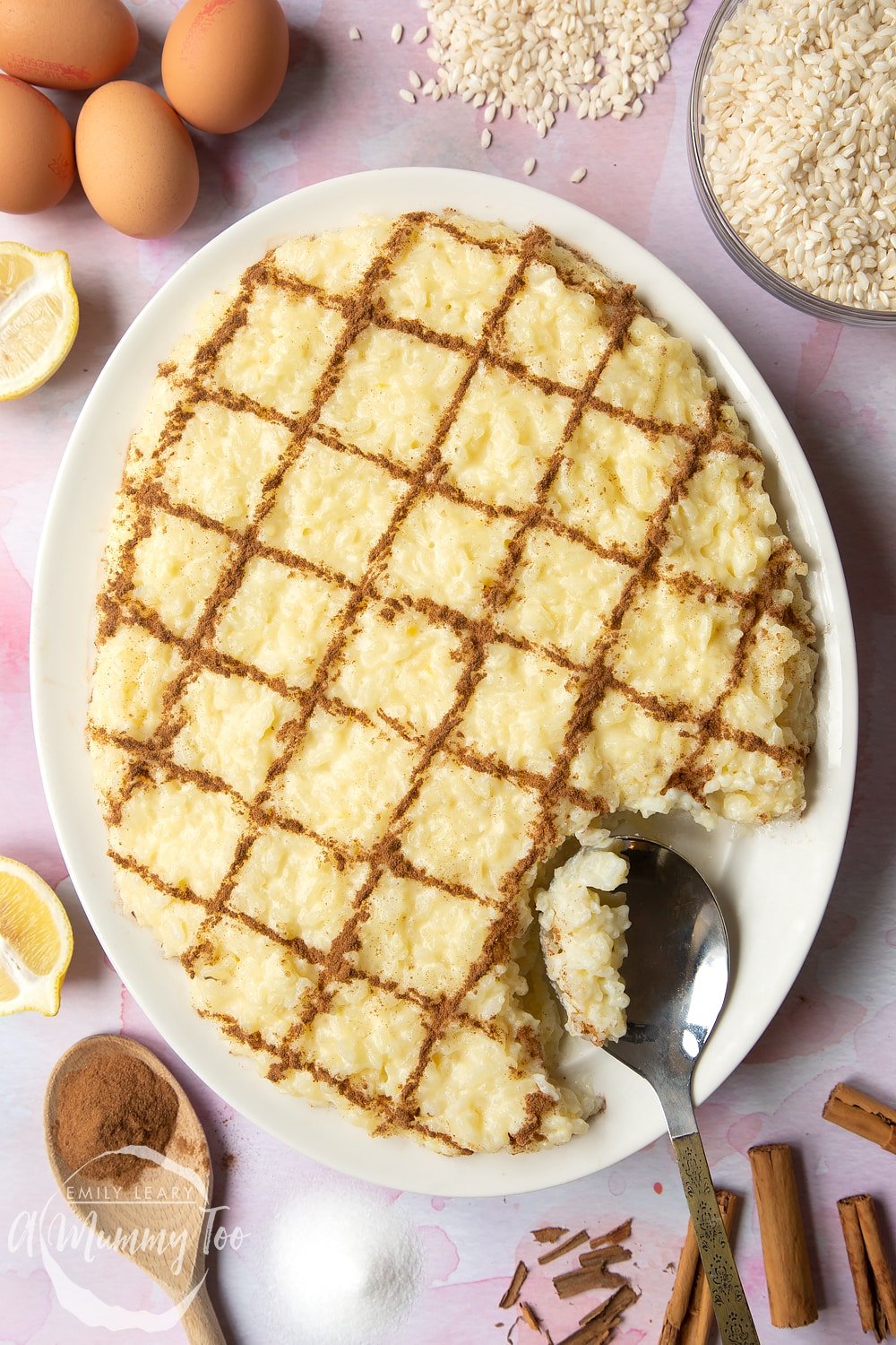 Arroz Doce (Portuguese rice pudding) on a large, white, oval-shaped plate. The rice pudding is decorated with ground cinnamon in a crosshatch pattern. A spoon rests on the plate.