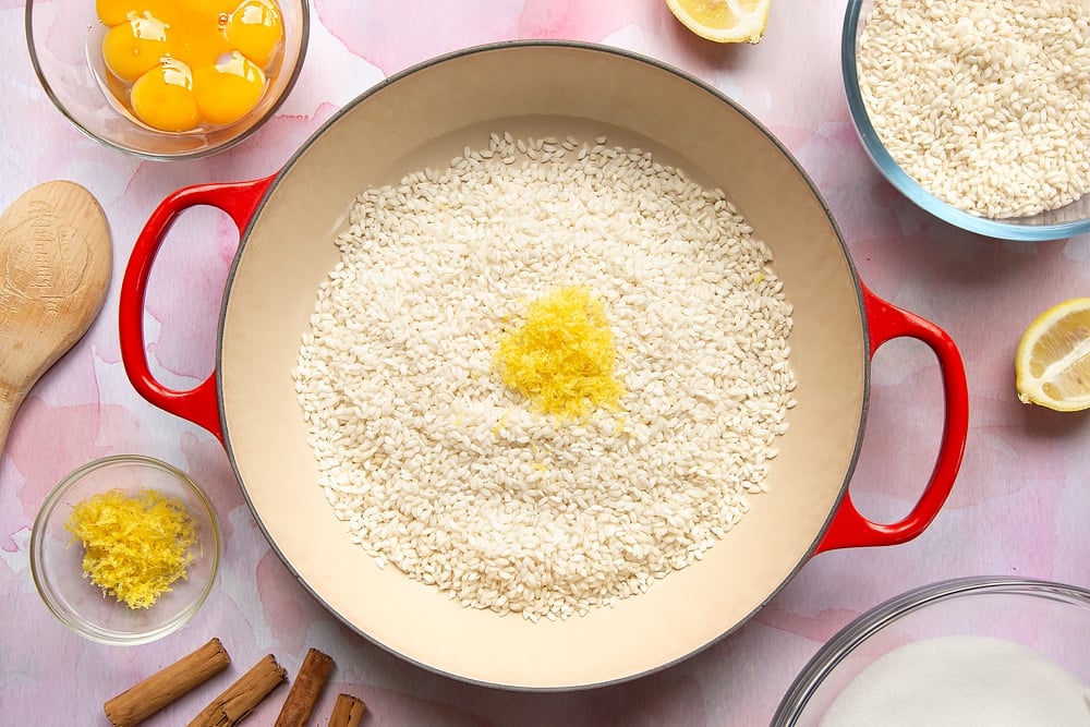 A pan filled with arborio rice and lemon zest. Around the pan is the ingredients to make Arroz Doce.